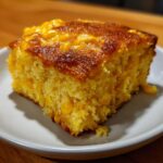 A close-up of a square slice of moist cornbread casserole with visible corn kernels, served on a white plate.