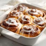 Close-up of six freshly baked small batch cinnamon rolls covered in thick white icing in a white baking dish.