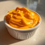 A close-up of rich, vibrant orange nacho cheese sauce with soft peaks in a small white ramekin.