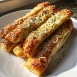 Close-up of several golden brown, soft breadsticks topped with melted cheese and dried herbs on a white plate.