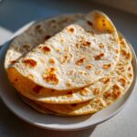 A close-up stack of freshly made, soft corn tortillas with golden-brown spots resting on a white plate.