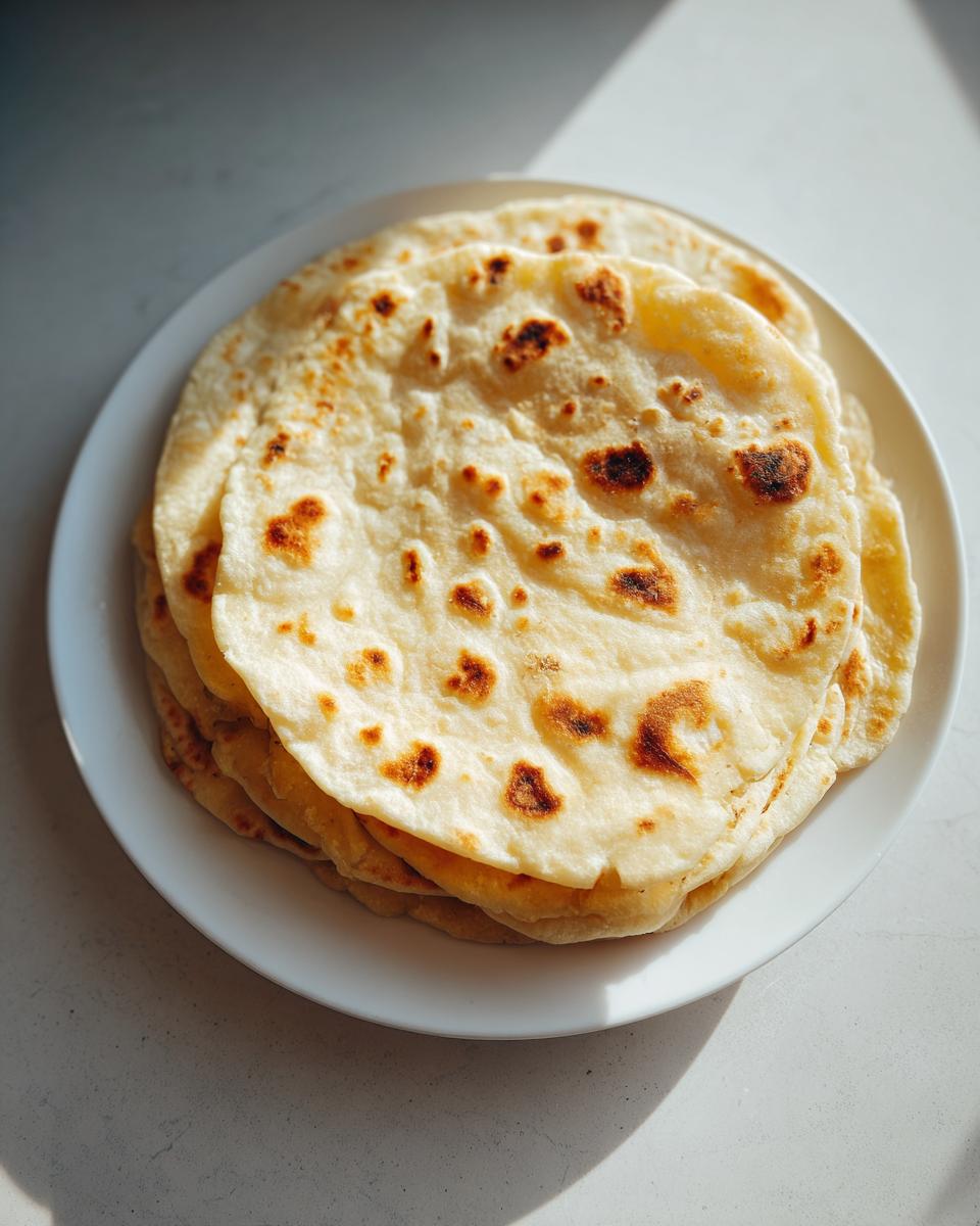 A stack of freshly made, soft corn tortillas with light brown charred spots resting on a white plate.