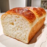 A freshly baked loaf of incredibly soft milk bread, showing a fluffy white interior and a shiny, golden-brown top crust.