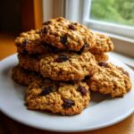A tempting stack of soft oatmeal cookies loaded with raisins resting on a white plate near a window.