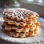 A tall stack of golden brown pizzelle cookies, featuring a delicate waffle pattern and heavily dusted with powdered sugar.