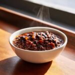 A close-up of a steaming white bowl filled with rich, dark black bean chili resting on a wooden surface.