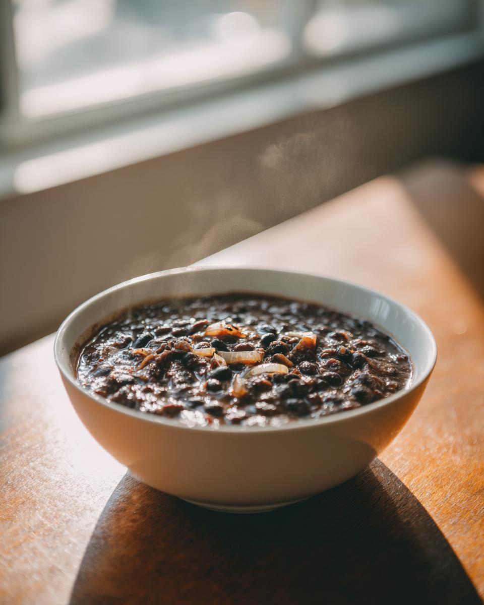 A steaming white bowl filled with rich, dark black bean chili topped with sautéed onions, set on a wooden table near a window.