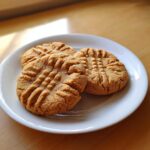 Three freshly baked peanut butter cookies with classic crosshatch fork marks, resting on a white plate.