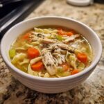 A close-up of a white bowl filled with homemade turkey soup, featuring shredded turkey, egg noodles, carrots, and celery.