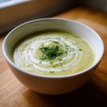 A close-up of a bowl of velvety leek soup, swirled with cream and topped with fresh dill.
