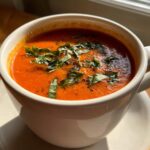 Close-up of a rich, orange-red tomato bisque topped with fresh basil leaves and cracked black pepper.