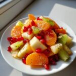 Close-up of a bright winter fruit salad featuring orange segments, kiwi chunks, apple pieces, and ruby pomegranate seeds.