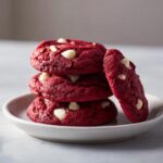 A stack of three vibrant red velvet cake mix cookies with white chocolate chips, leaning against one cookie on a small white plate.