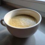 A white mug filled with creamy, frothy coffee sitting on a windowsill, unrelated to roasted cauliflower soup.