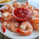 A white plate featuring perfectly cooked shrimp cocktail surrounding a small glass bowl of bright red cocktail sauce.