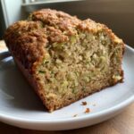A close-up of a thick slice of moist zucchini banana bread showing green shreds of zucchini in the crumb.