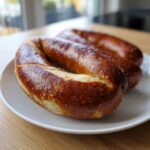 Three freshly cooked pretzel dogs with a dark, shiny crust and coarse salt, resting on a white plate.