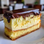 A cross-section slice of boston cream pie cheesecake showing layers of yellow cake, vanilla cream filling, and chocolate ganache topping.