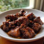 Close-up of rich, dark brown stew chicken pieces coated in thick sauce served in a white bowl.