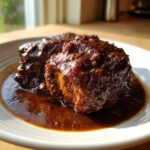 Close-up of rich, dark brown stew chicken Jamaican recipe pieces covered in thick sauce on a white plate.