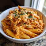 A close-up of creamy, shredded chicken in a rich sauce mixed with penne pasta, topped with melted cheese and parsley, representing cajun chicken pasta crockpot.