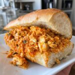 A messy, saucy Cajun chicken sloppy joes filling served inside a split, crusty white roll on a white plate.