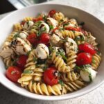 A close-up of caprese chicken pasta salad featuring fusilli pasta, cherry tomatoes, mozzarella balls, and chicken chunks drizzled with balsamic glaze.