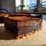 A close-up, sunlit slice of rich chocolate cheesecake bars showing a graham cracker crust and two layers of dark chocolate filling.