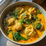 Close-up of a bowl of vibrant yellow coconut curry soup with dumplings and wilted spinach, garnished with cilantro.