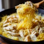 A wooden spoon lifts strands of spaghetti squash mixed with creamy sauce and seasoned chicken pieces in a skillet, showcasing the chicken spaghetti squash.