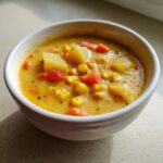 Close-up of a creamy, yellow corn chowder soup with chunks of potato, corn, and carrots in a white bowl.