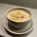 A white bowl filled with creamy crab and corn chowder, showing chunks of crab meat, bright yellow corn, and diced carrots.