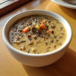 A close-up of a white bowl filled with creamy wild rice soup, showing dark wild rice grains and diced carrots.