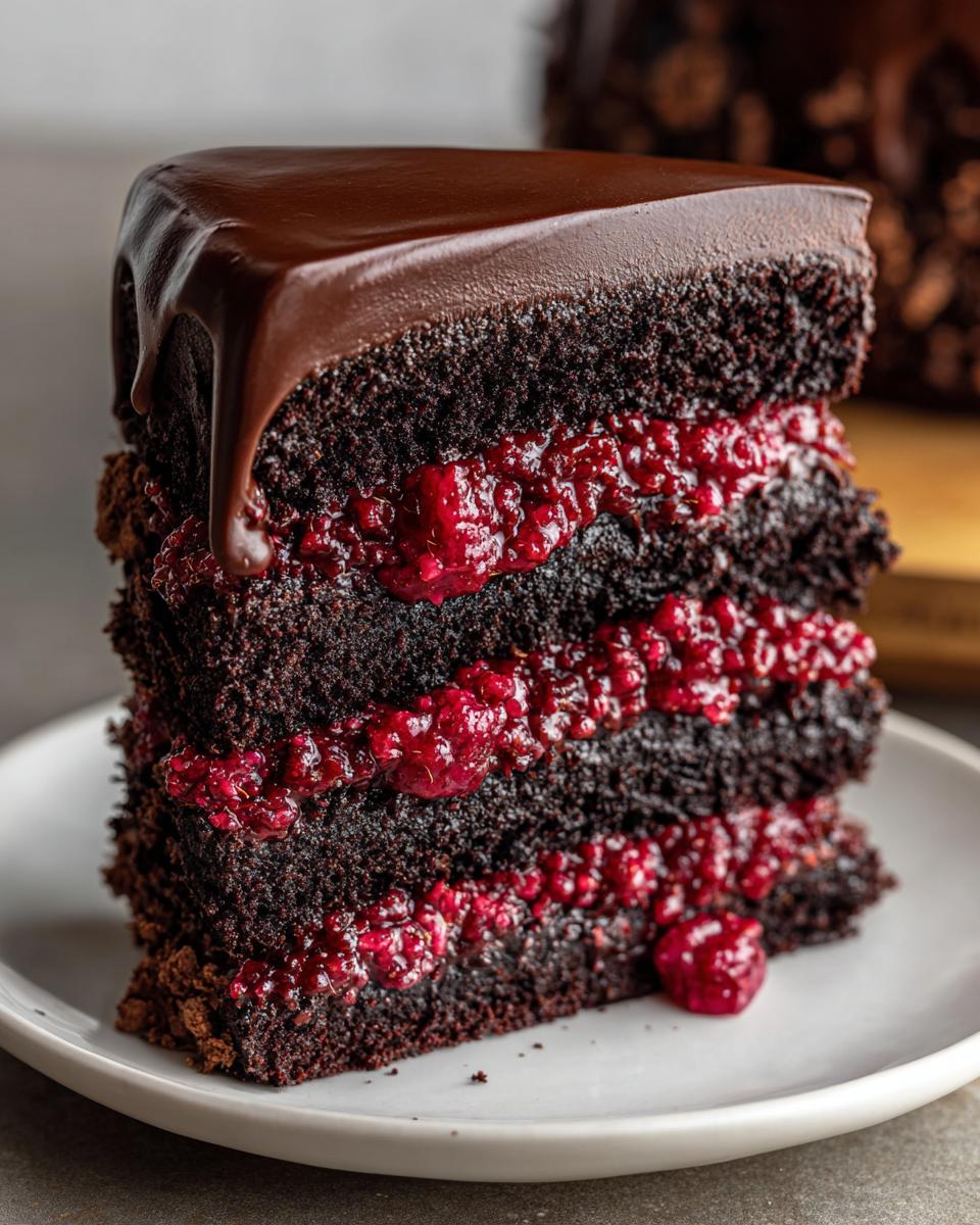 Close-up of a decadent slice of dark chocolate raspberry cake with three layers of filling and a shiny chocolate ganache drip.