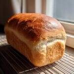 A golden-brown, freshly baked yeast bread loaf cooling on a wire rack near a sunlit window.