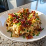 A plate of loaded smashed potato salad, topped generously with creamy sauce, shredded cheese, bacon bits, and fresh chives.