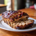 A moist stove top stuffing meatloaf, sliced and covered in a dark, shiny glaze, resting on a white plate.