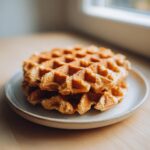 Close-up of a stack of golden brown oatmeal cottage cheese waffles on a speckled white plate.