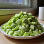 A mound of bright green, powdered sugar-dusted pistachio puppy chow chex mix piled high on a white plate.
