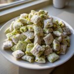 A generous pile of sweet, powdered sugar-dusted pistachio puppy chow mix featuring bright green pistachio pieces.