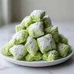 A mound of bright green pistachio puppy chow generously dusted with powdered sugar, served on a white plate.