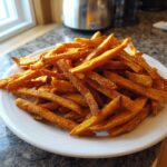 A large pile of crispy, orange sweet potato fries heavily seasoned with dark spices, served on a white plate.