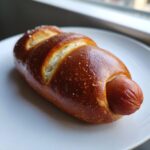 A single, freshly baked sourdough discard pretzel dog with a shiny, dark brown crust and coarse salt, resting on a white plate.