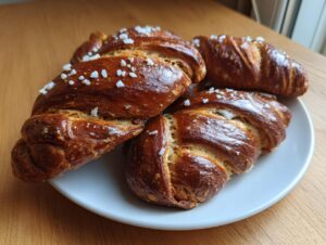 Four golden-brown, twisted sourdough pretzel dogs topped with coarse salt resting on a white plate.