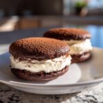 Two rich, dark chocolate tiramisu cookies filled with creamy white frosting and dusted with cocoa powder.