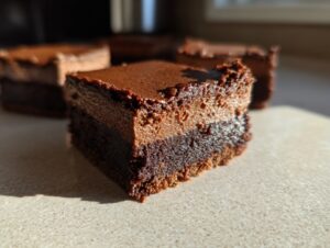 A close-up, sunlit view of a square chocolate cheesecake bars piece showing a dense brownie base and a lighter chocolate layer.