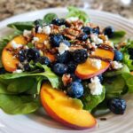 A vibrant close-up of a blueberry peach feta salad featuring spinach, fresh peach slices, blueberries, feta cheese, pecans, and a balsamic glaze.