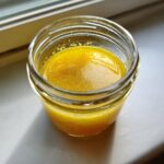 A small glass jar filled with bright yellow lemon vinaigrette salad dressing, resting on a white surface near a window.