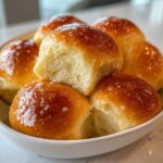 Close-up of soft, golden-brown dinner rolls brushed with melted butter, piled in a white bowl.