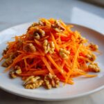 A vibrant mound of shredded carrots topped with whole walnuts, representing a carrot apple walnut salad.